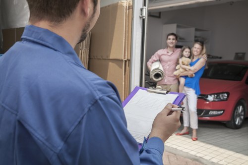 A man wearing a blue shirt holds a clipboard and pen, standing outside a residential garage in a residential area near Bermondsey. Inside the open garage, there are several cardboard moving boxes stacked on the driveway, which has a textured paving surface. To the right, a red vehicle is parked close to the garage entrance, partially visible with a sleek, modern design. In front of the garage, a family of three, including a man, woman, and a young child, are standing and smiling towards the man with the clipboard. The woman is holding the child, and both are dressed casually; the man is in a light brown shirt, and the woman is in a blue top. The background shows part of the house’s exterior wall with a neutral finish, and the scene is set during daylight with natural lighting illuminating the area. The image captures a moment of coordination involving house removals or moving services, reflective of Bermondsey Man with Van’s professional handling of home relocations.