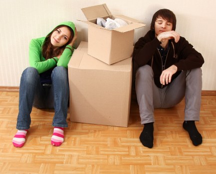 A young woman with long brown hair, wearing a green hoodie, blue jeans, and pink-and-white striped socks, sits on a wooden floor beside a large cardboard moving box with an open lid. She leans her head on her hands and appears to be resting or waiting. To her right, a young man with dark hair, dressed in a black hoodie, grey jeans, and black shoes, sits on the floor with his arms resting on his knees, appearing contemplative or tired. Between them and behind the woman is a smaller cardboard box filled with crumpled packing paper. The background features a plain, light-colored wall typical of a residential interior, indicating an indoor setting during a house move or packing process. The scene is well-lit, emphasizing the casual atmosphere of a household during relocation, with an emphasis on furniture, packing materials, and personal belongings typical of house removals services in Bermondsey or nearby areas.