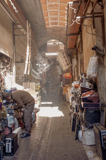 A man with a walking cane, dressed in a dark jacket and light trousers, stands inside a market stall during a home relocation or packing process, viewed from behind. The stall is filled with various i