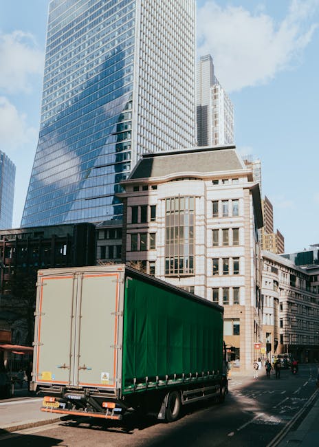 A large green box trailer attached to a commercial vehicle is parked on a city street surrounded by tall modern office buildings with reflective glass facades and older architectural styles. The trail